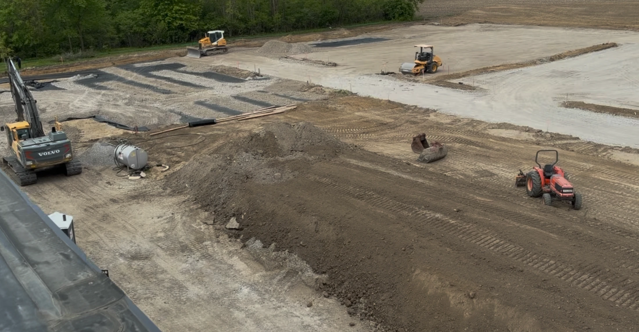 An aerial view of a construction site with various heavy machinery, including an excavator, bulldozer, and tractor, preparing a large dirt and gravel area.