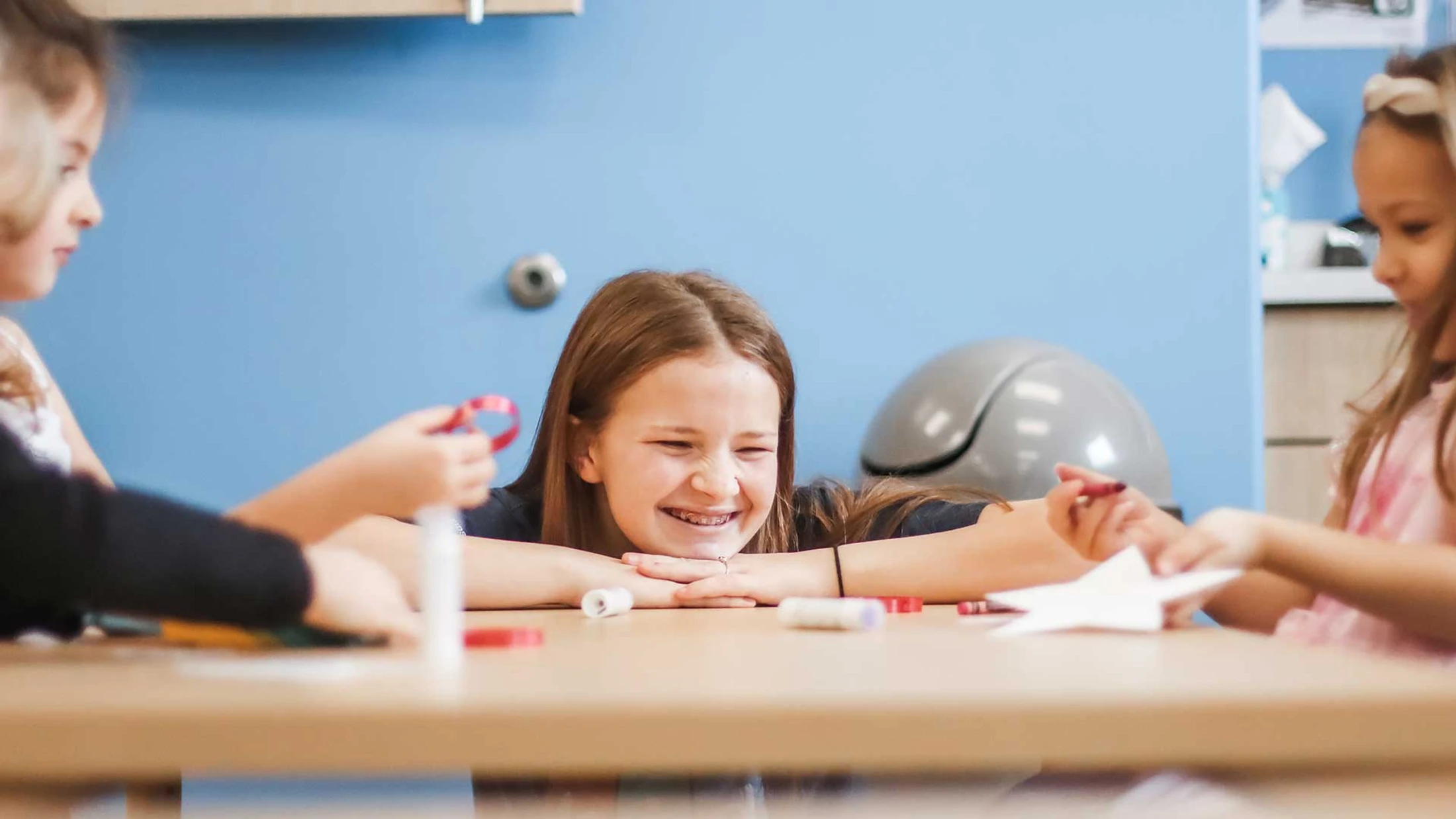 A young girl with braces smiles happily, resting her chin on her hands at a table with two other girls during an arts and crafts session.