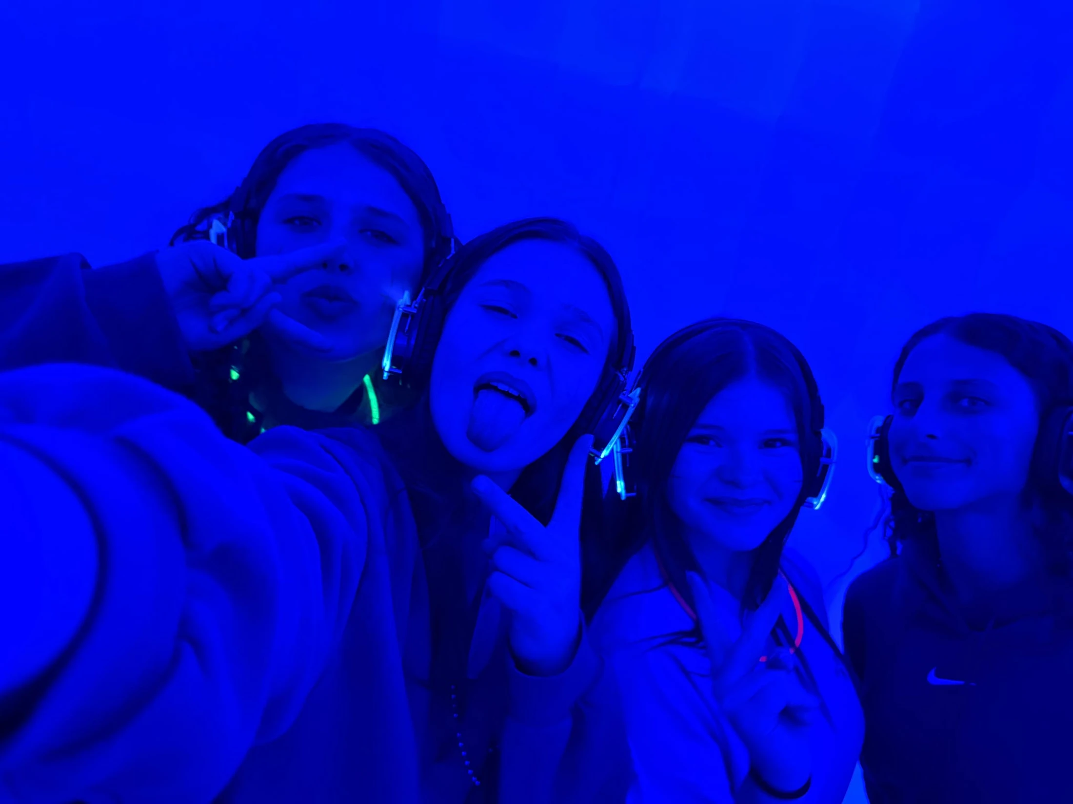 Four young women wear headphones and pose for a selfie under intense blue light, making playful faces and peace signs at a party.