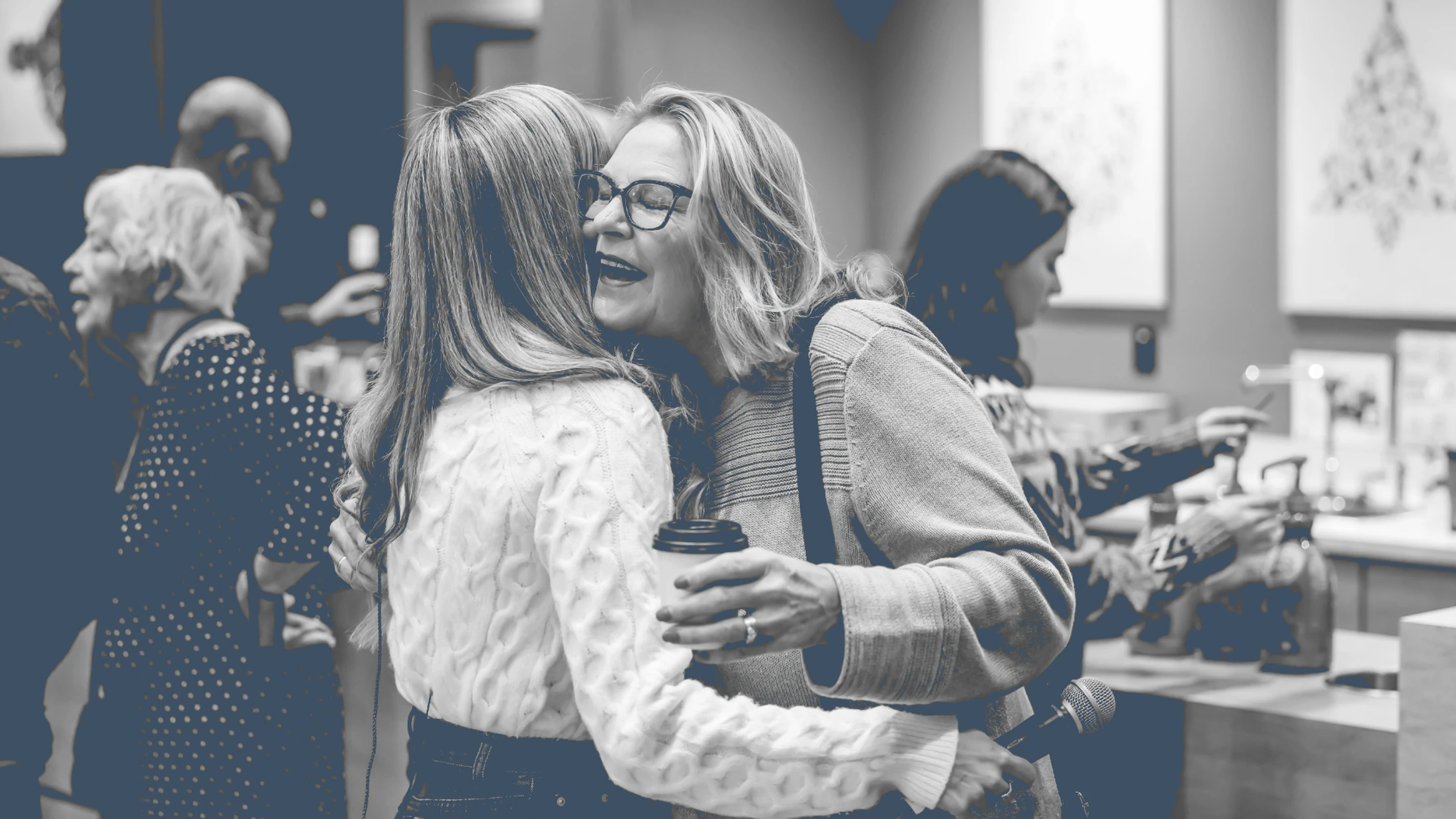 A black and white photo of two women hugging. One woman, wearing glasses and holding a coffee cup and microphone, is laughing as she embraces the other.