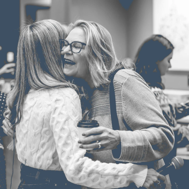 A black and white photo of two women hugging. One woman, wearing glasses and holding a coffee cup and microphone, is laughing as she embraces the other.
