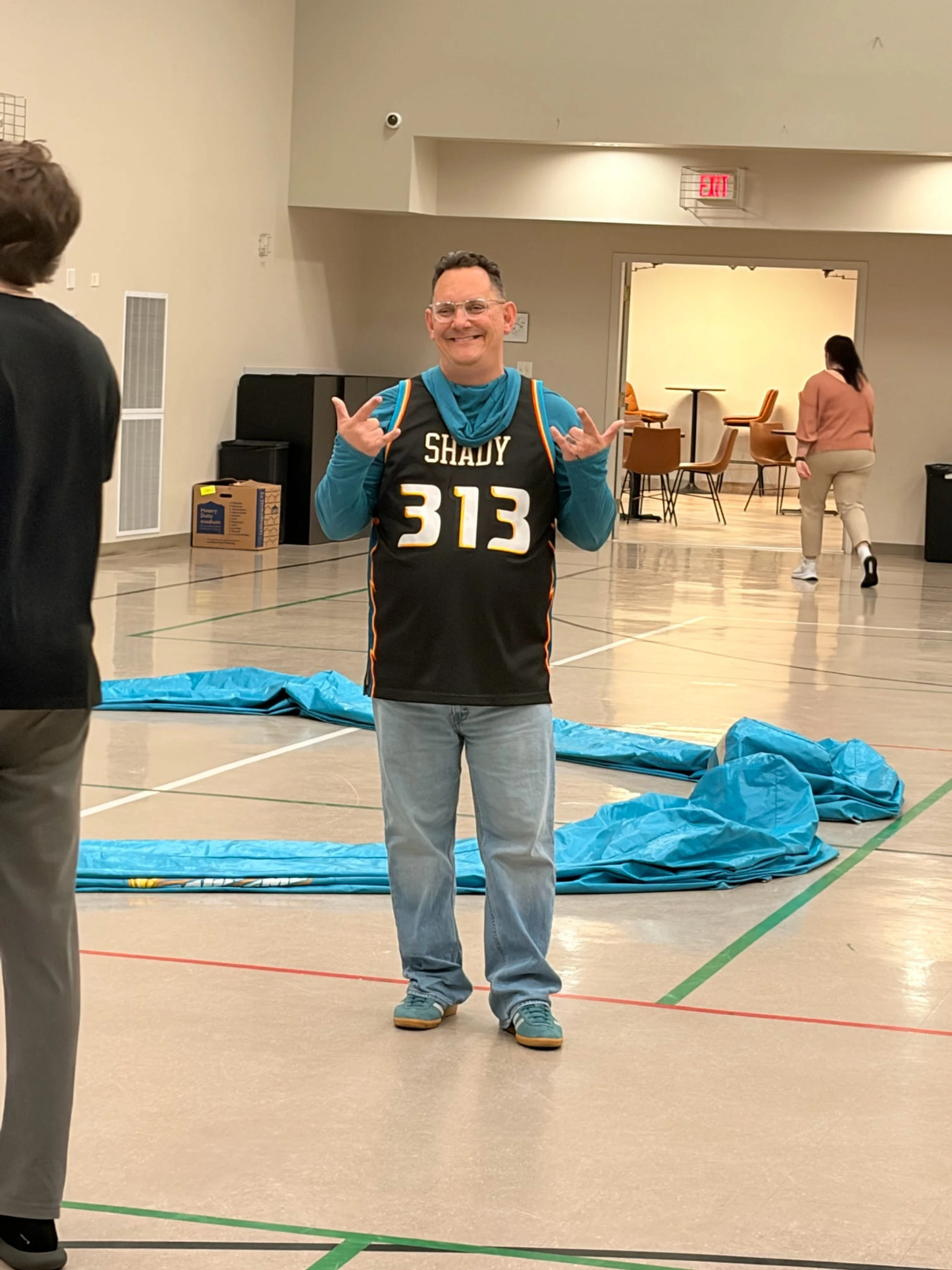 A smiling man in glasses and a black 'Shady 313' basketball jersey makes a hand gesture while standing in the middle of a gymnasium.