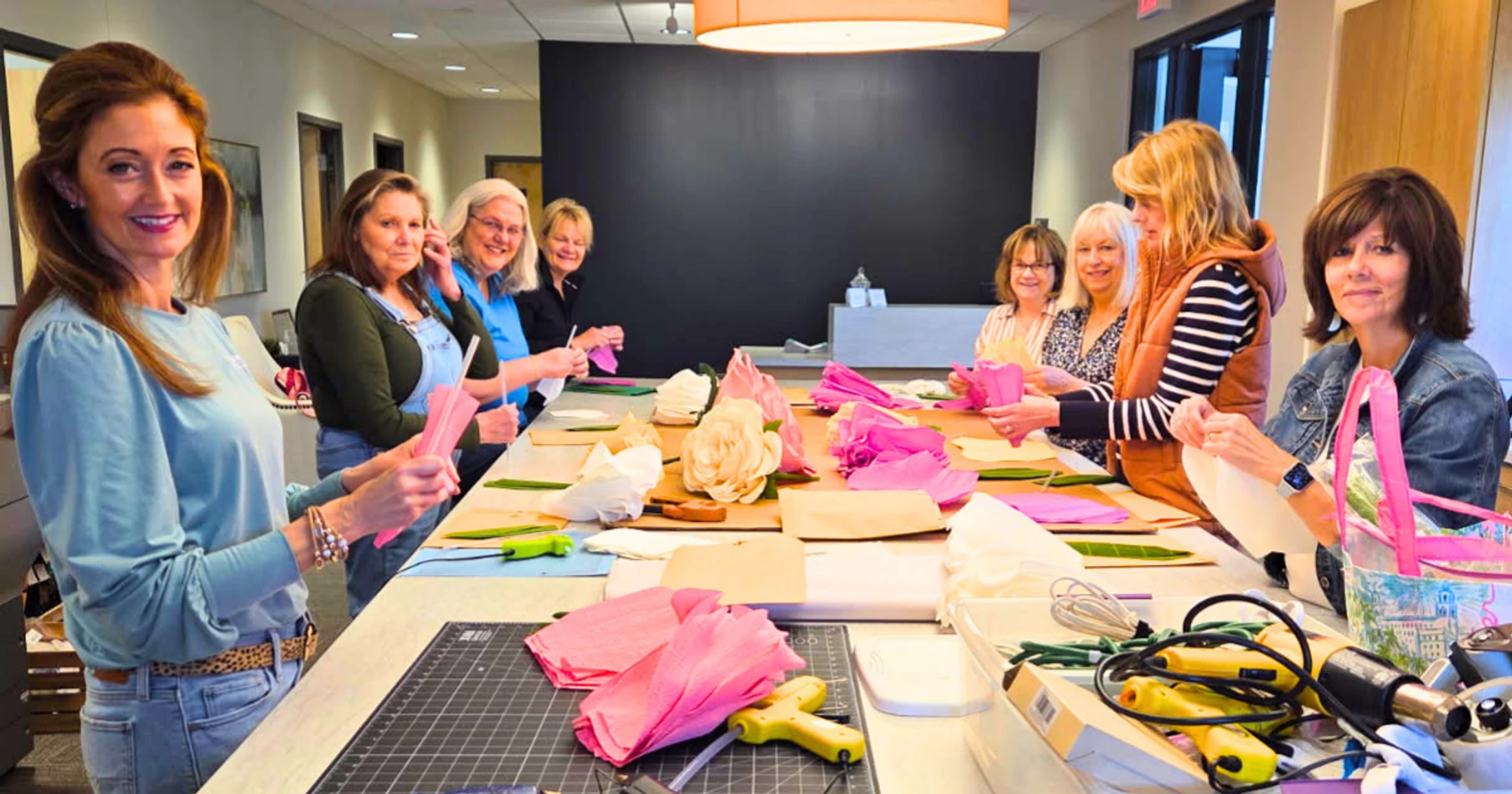 A group of smiling women gathered around a long table in a workshop, learning to make pink and white paper flowers with various craft supplies.