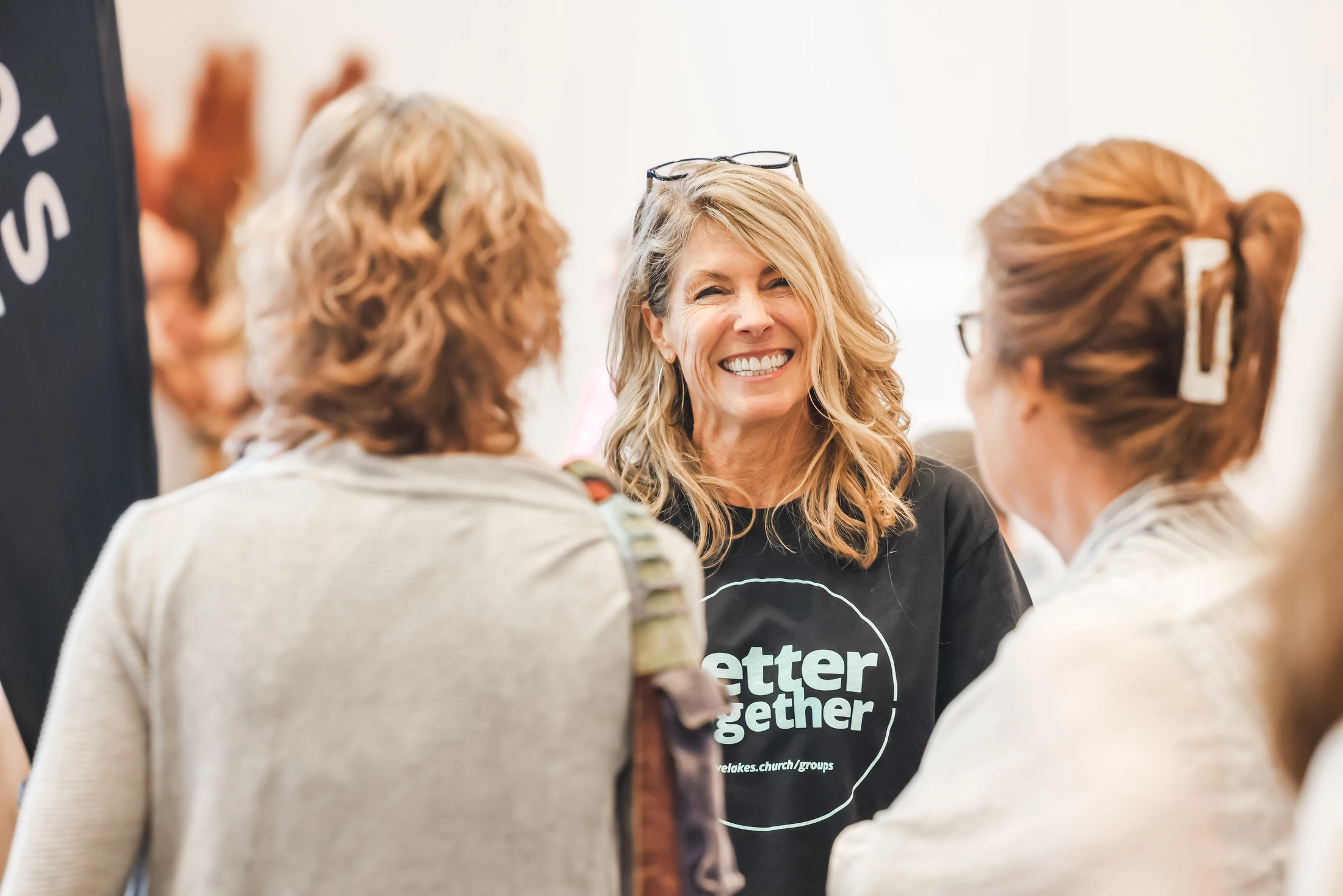 A smiling blonde woman in a black 'better together' t-shirt talks with two other women, who are seen from behind, in a brightly lit room.