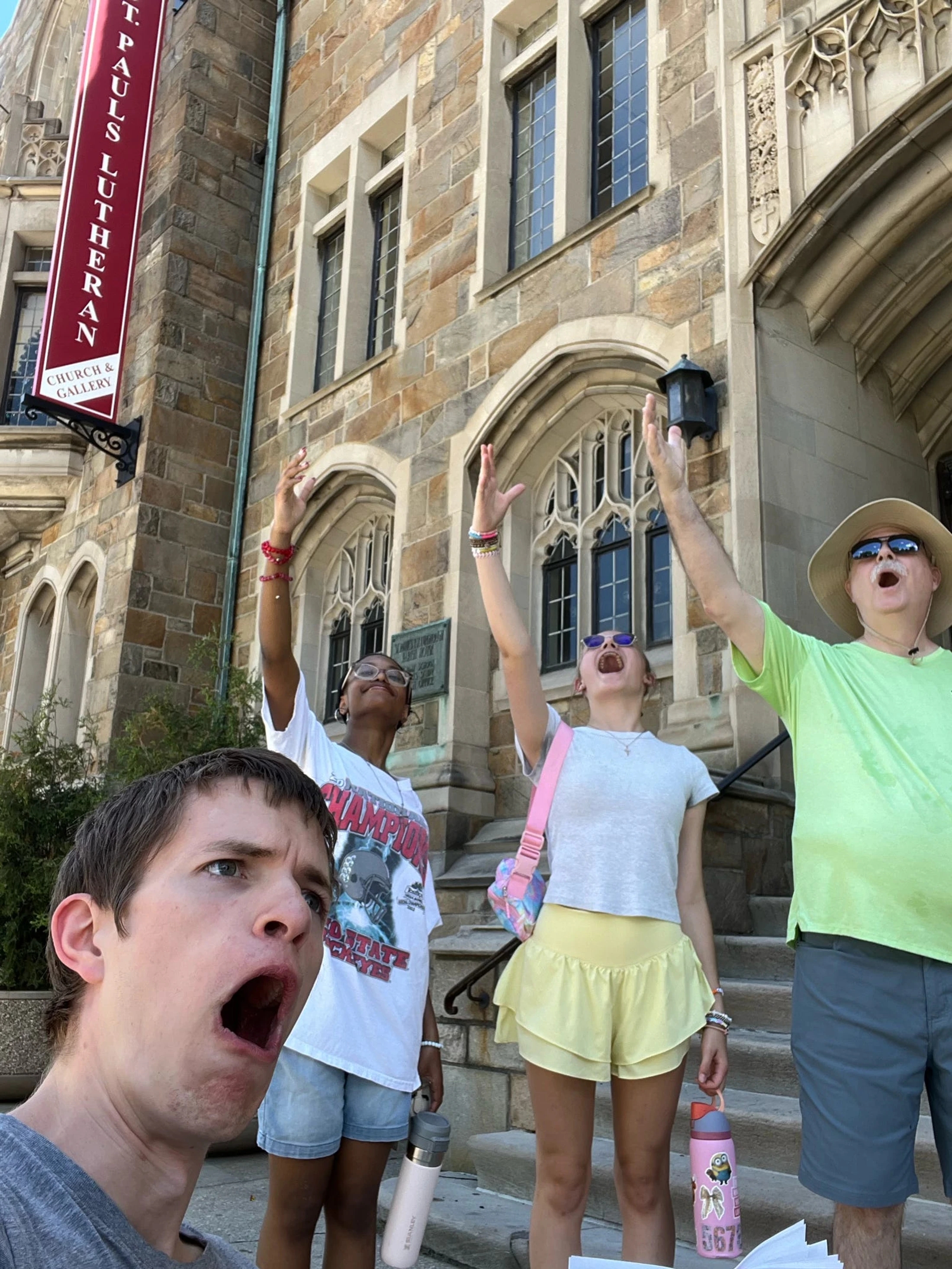 Four people with exaggerated, open-mouthed expressions pose for a selfie on the steps of a stone church, three with their arms raised.