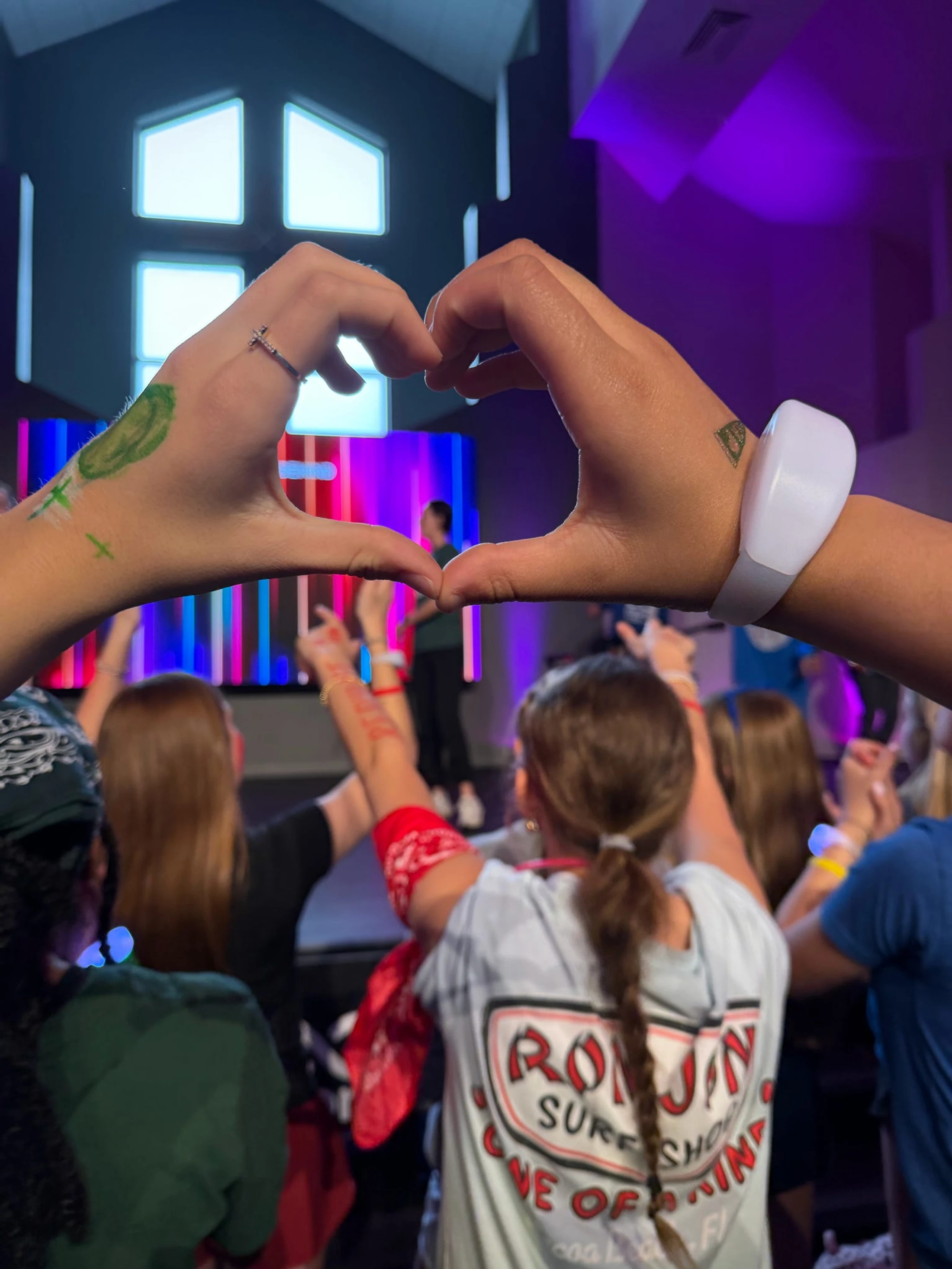 A close-up of two hands making a heart shape, overlooking a crowd of young people worshipping in front of a brightly lit stage with purple lights.