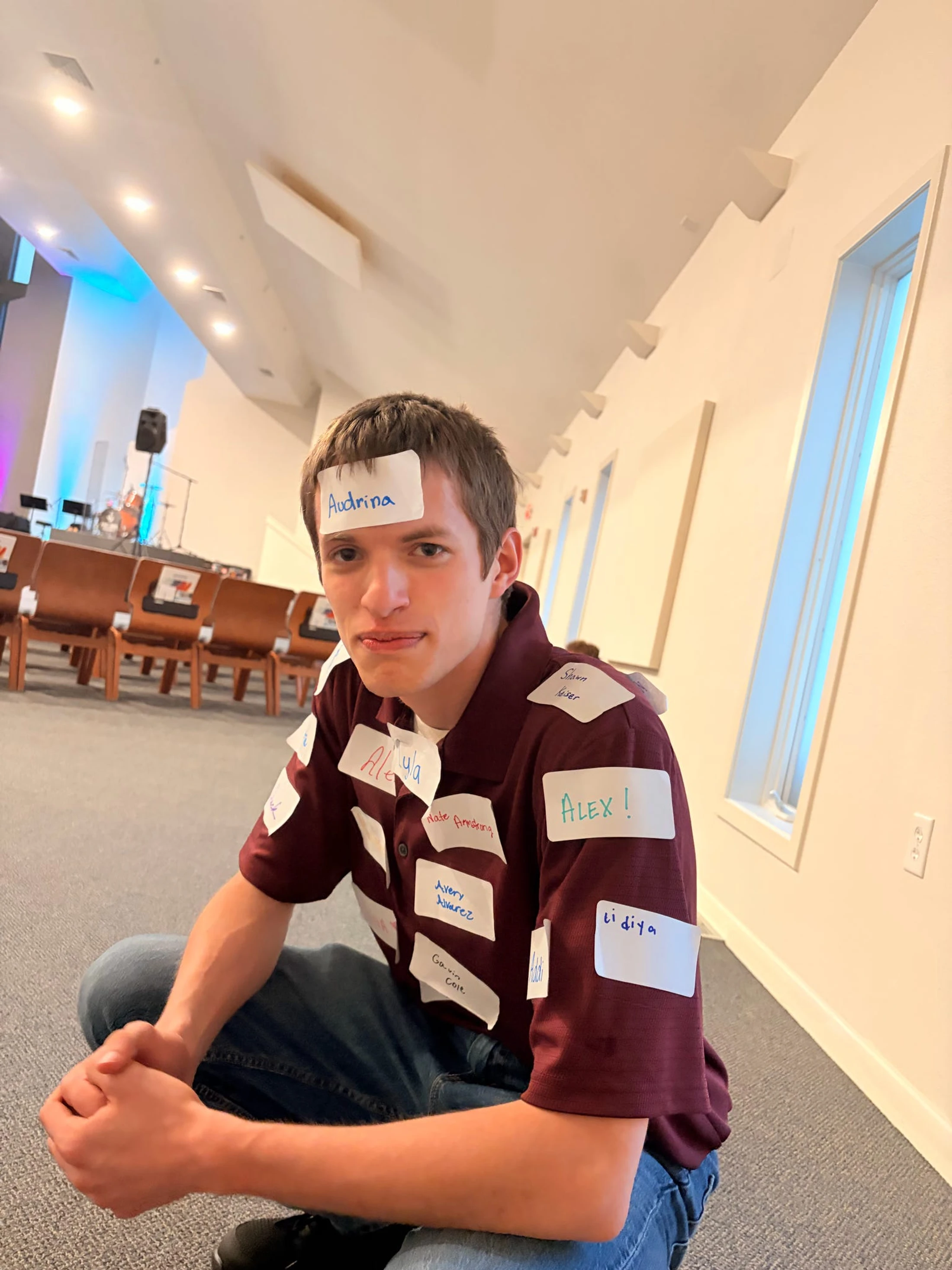 A young man sits cross-legged on a carpeted floor, looking at the camera with his shirt and forehead covered in white name tag stickers.