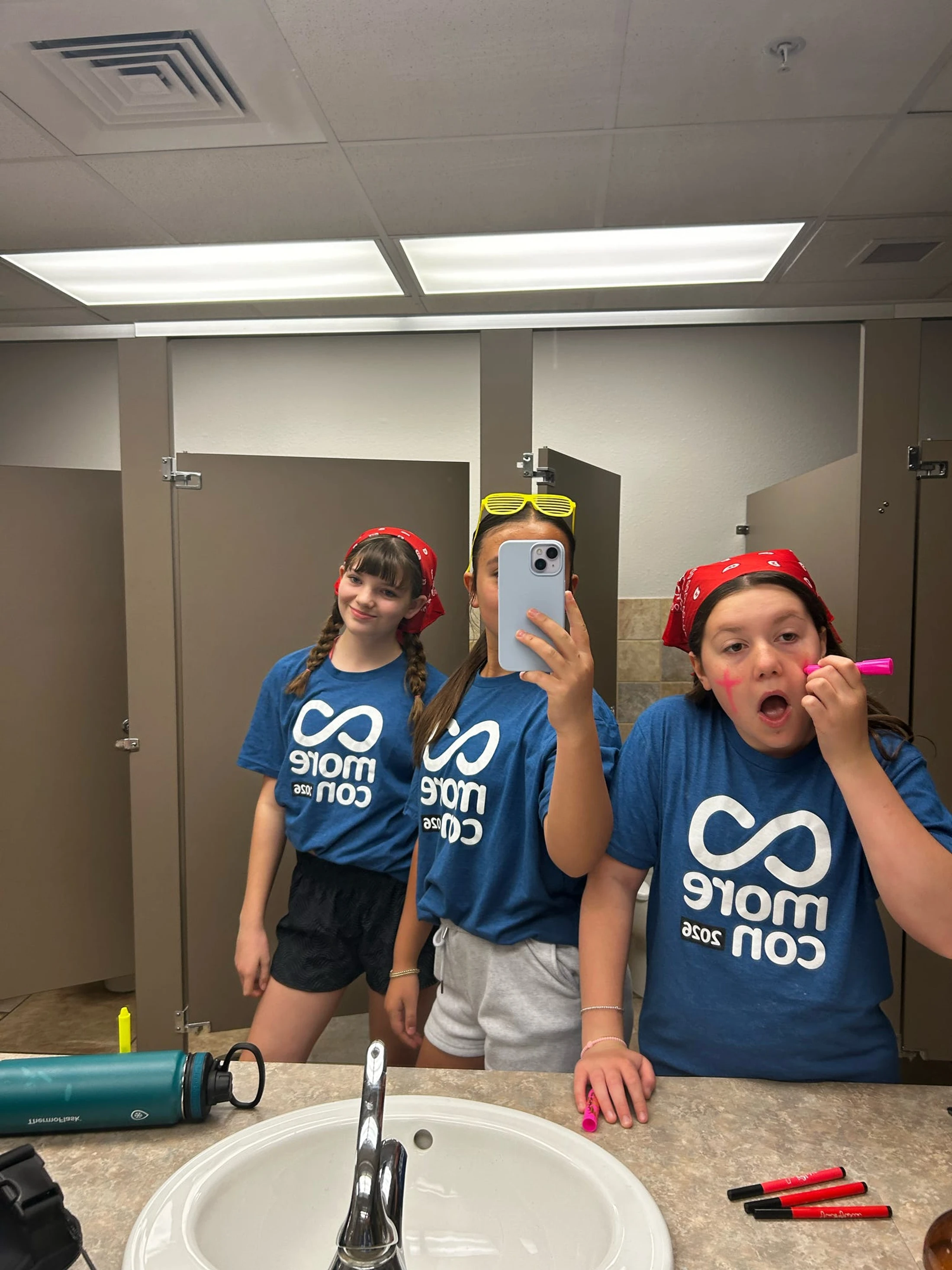 Three young girls in matching blue t-shirts take a selfie in a bathroom mirror. One girl holds the phone, while another playfully applies pink face paint.