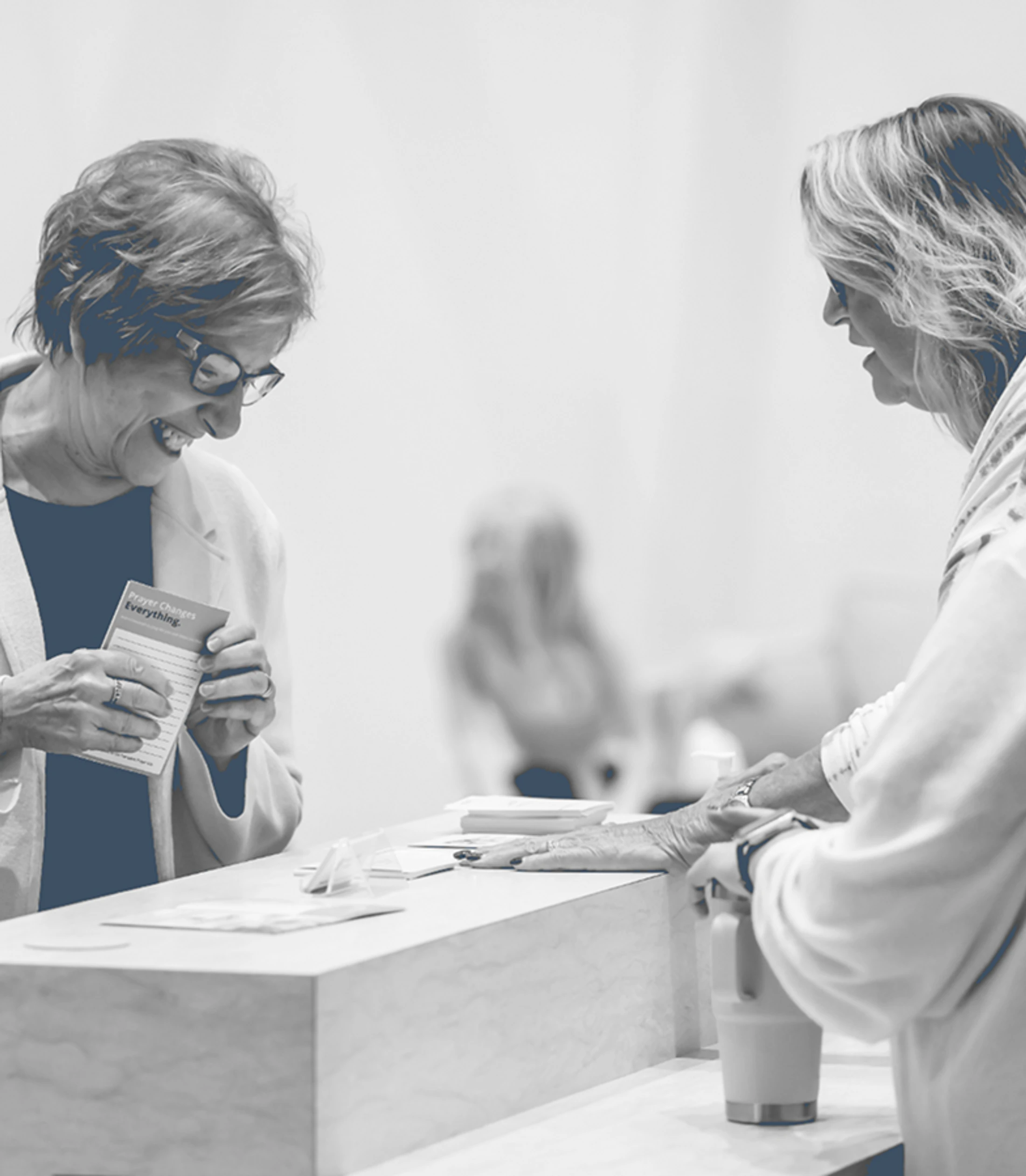 A black and white photo of two women at a counter. One woman smiles while holding a small booklet that says 'Prayer Changes Everything.'