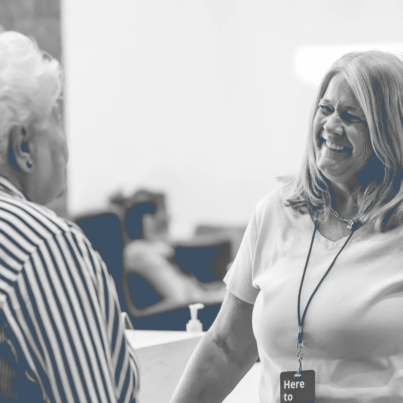 A black and white photo with a blue tint of a smiling woman with a lanyard talking warmly to another woman who has her back to the camera.