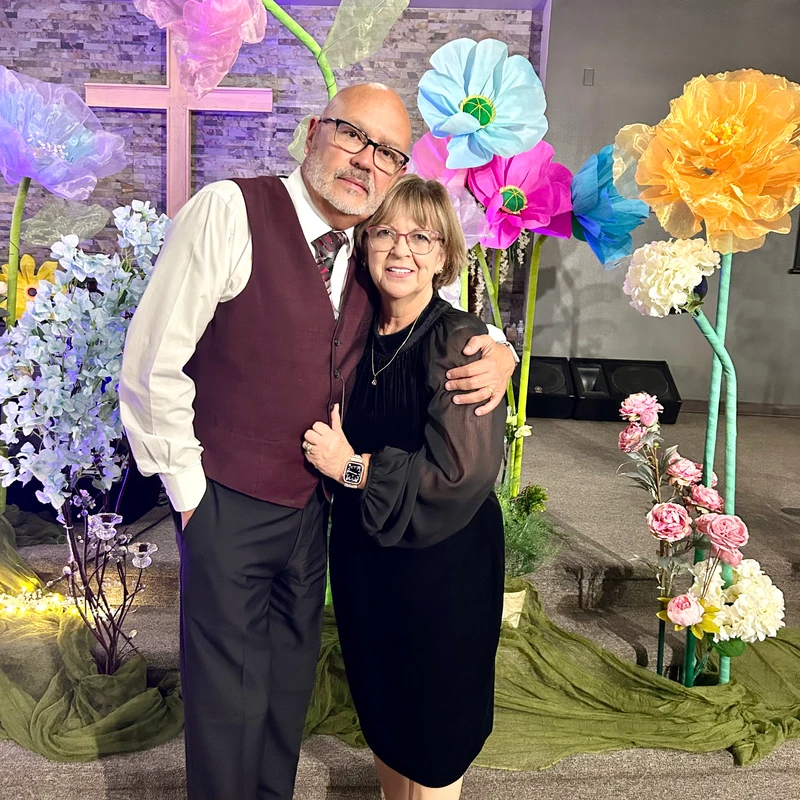 A man and woman dressed up, embracing and smiling for the camera. They stand in front of a backdrop with a cross and large, colorful artificial flowers.