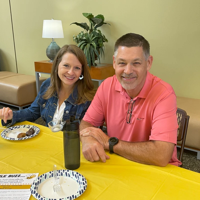 A man in a pink polo shirt and a woman in a denim jacket sit side-by-side at a table with a yellow tablecloth, smiling warmly at the camera.