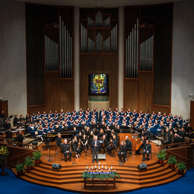 A wide shot of a large choir in blue robes and an orchestra performing on a tiered wooden stage, with a large pipe organ and stained glass window behind them.