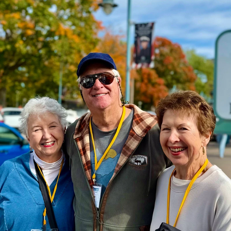 A man flanked by two women, all older adults, smile happily for a photo outdoors. The background shows trees with fall foliage and a partial sign.