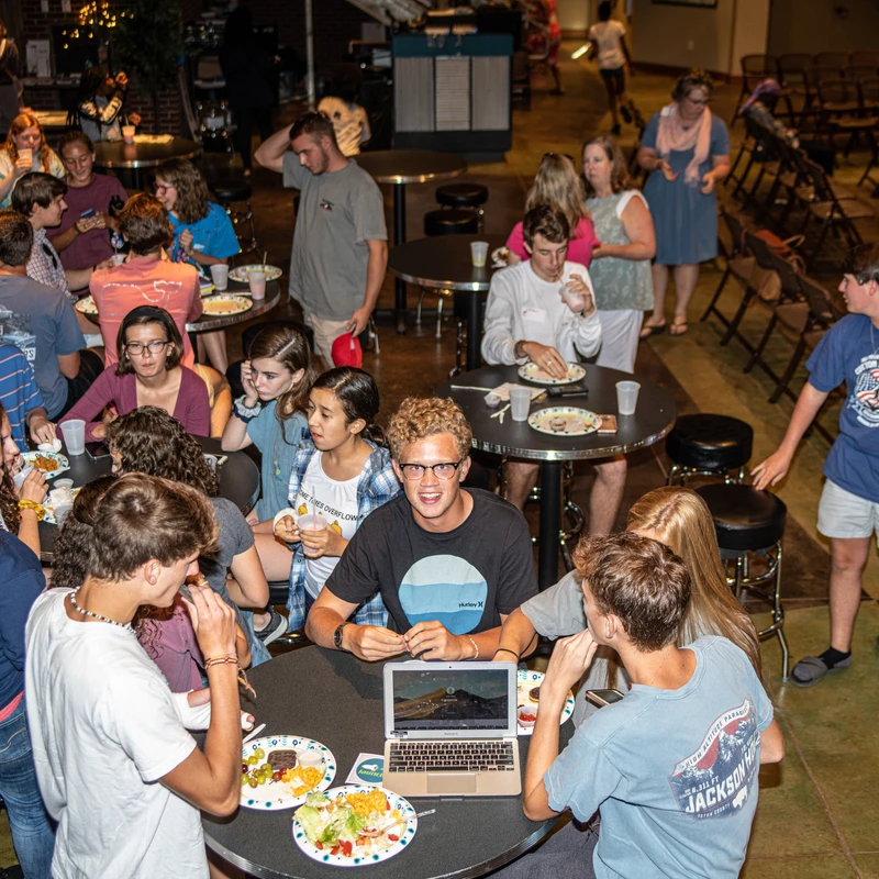 A large group of teenagers socializes and eats at round tables in a large hall. A boy in the center smiles at the camera with a laptop open before him.