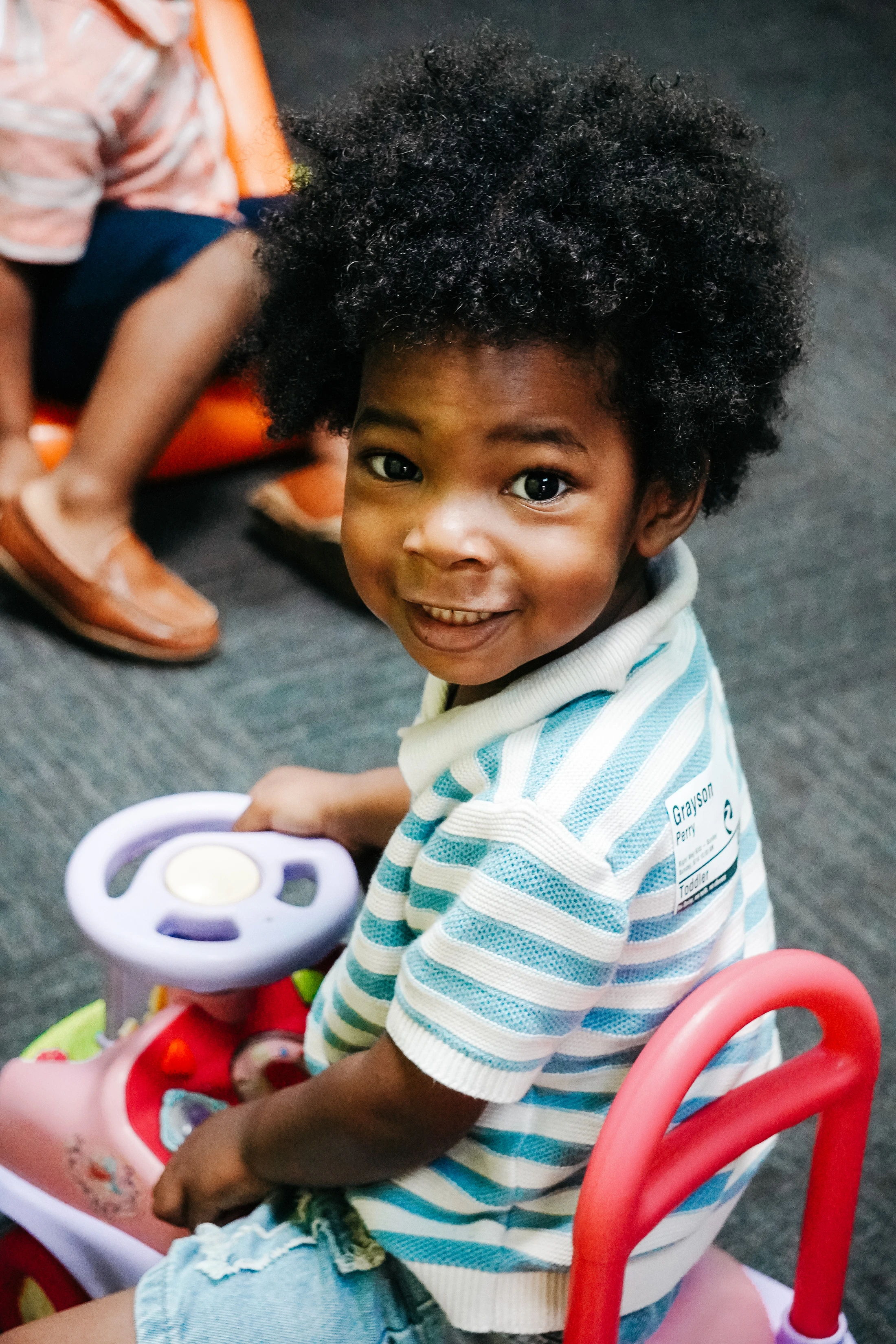 A young Black toddler with an afro hairstyle smiles at the camera while sitting in a colorful ride-on toy car.