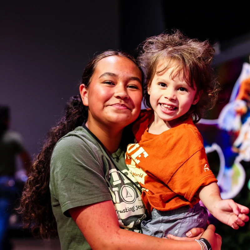 A young woman with long curly hair smiles while holding a small, happy child. They are both looking directly at the camera in a dimly lit room.