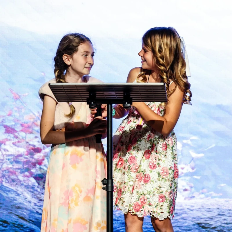 Two young girls in floral dresses stand together at a music stand on a brightly lit stage, smiling at each other during a performance.