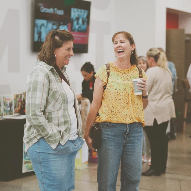 Two women stand together in a well-lit room, laughing. One wears a plaid shirt and denim skirt, the other a yellow top and jeans while holding a coffee cup.