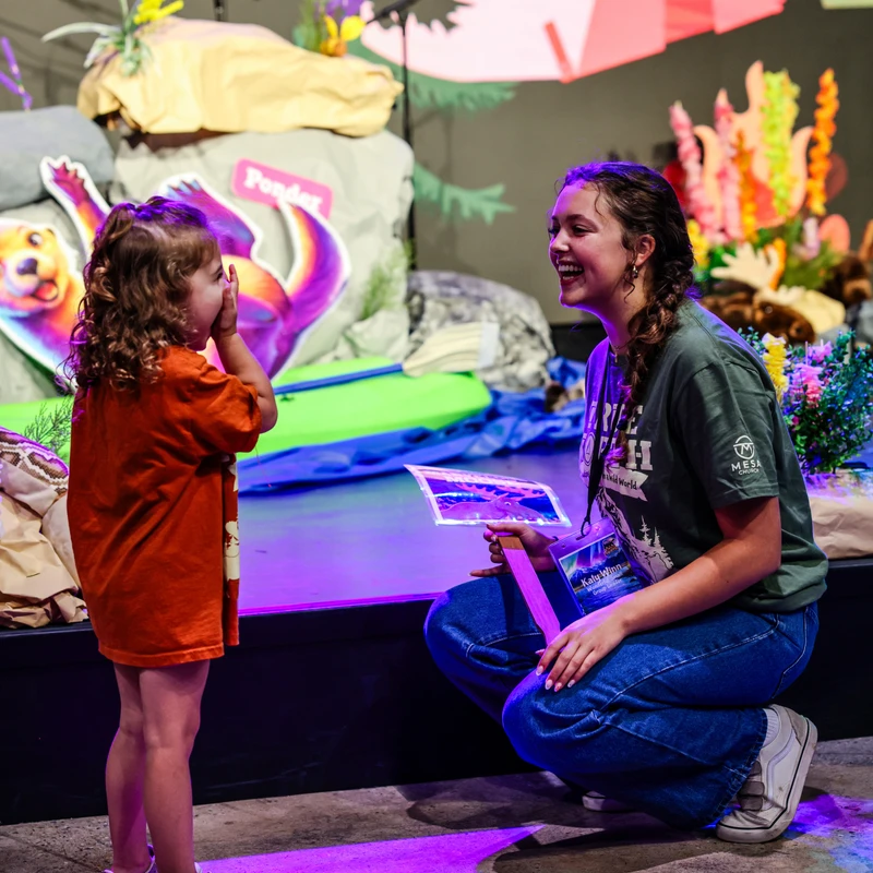 A young woman in a green shirt kneels and smiles at a small girl in an orange shirt, who covers her mouth and laughs on a purple-lit stage with fun decorations.