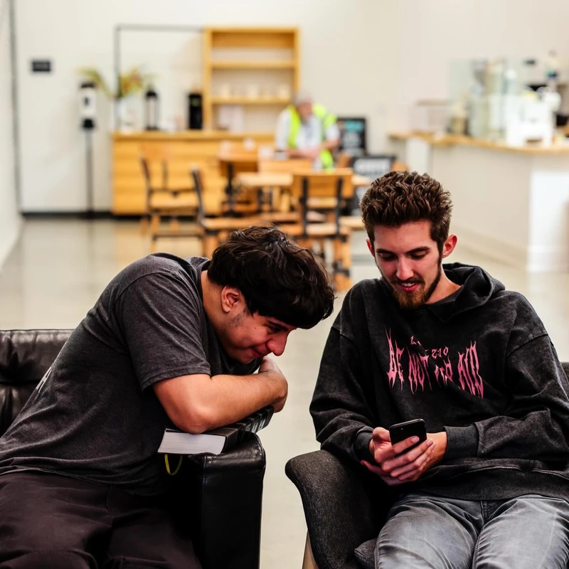 Two young men are sitting in chairs in a modern common area, smiling as they look together at something on a smartphone held by one of them.