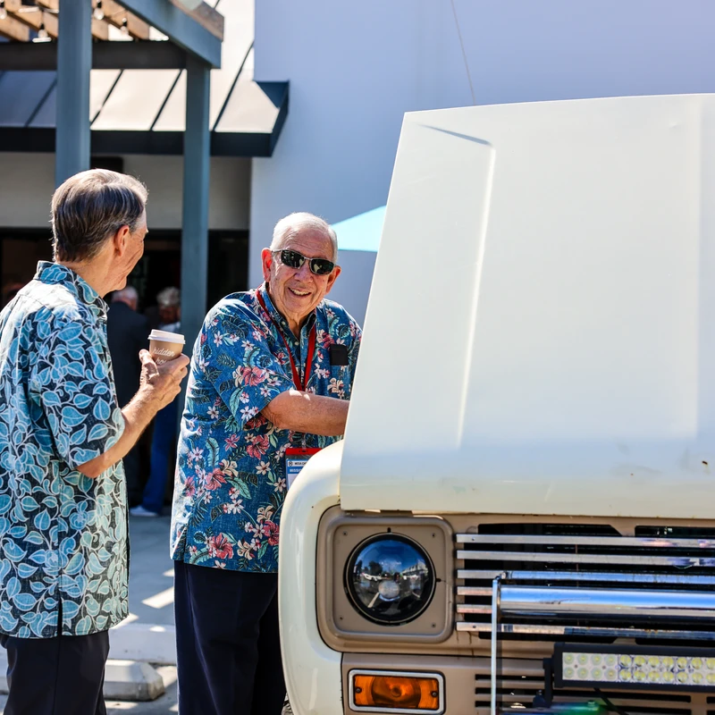 Two older men in colorful shirts chat by a vintage white off-road truck with its hood open. One man in sunglasses smiles warmly at the camera.
