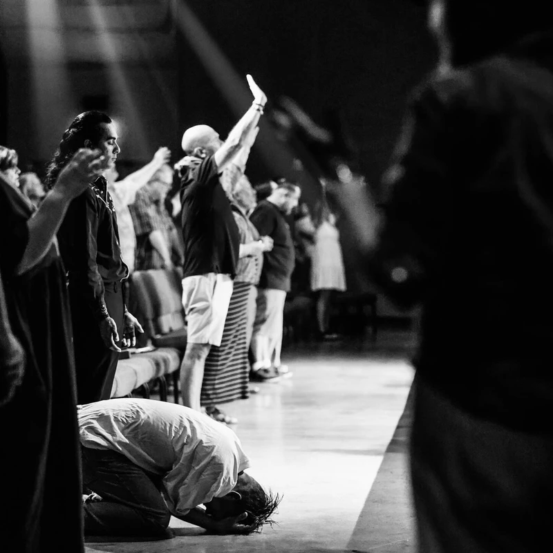 A black and white photo of a congregation worshiping. Some people stand with hands raised while one person kneels prostrate on the floor in the foreground.