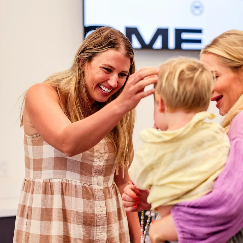 A smiling woman in a checkered dress gently touches the hair of a small blond child being held by another woman during an indoor event.