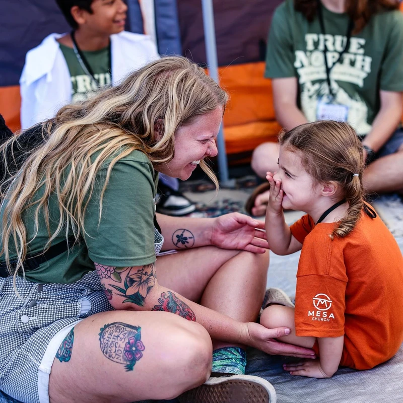 A smiling woman with tattoos sits on a tarp, laughing with a little girl in an orange shirt who is giggling with her hand over her mouth.