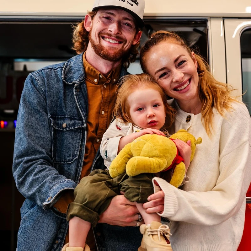 A happy young couple smiles while holding their toddler. The man wears a 'Good Dad' hat, and the child holds a Winnie the Pooh stuffed animal.