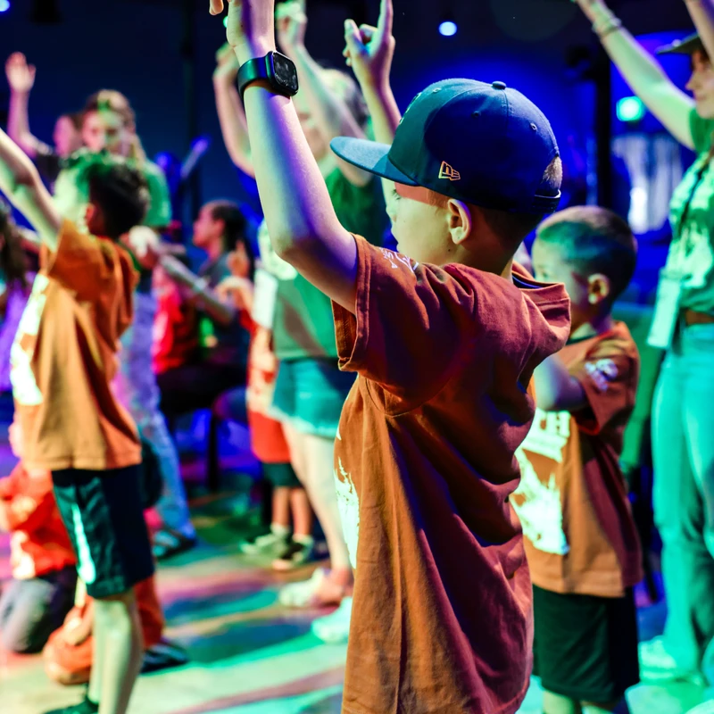 A young boy in a backward cap raises his finger to the sky, surrounded by other children doing the same in a room with colorful blue and green lights.