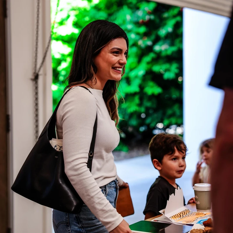 A smiling woman with dark hair stands next to a table where a young boy is sitting. The background is bright with greenery visible through a doorway.