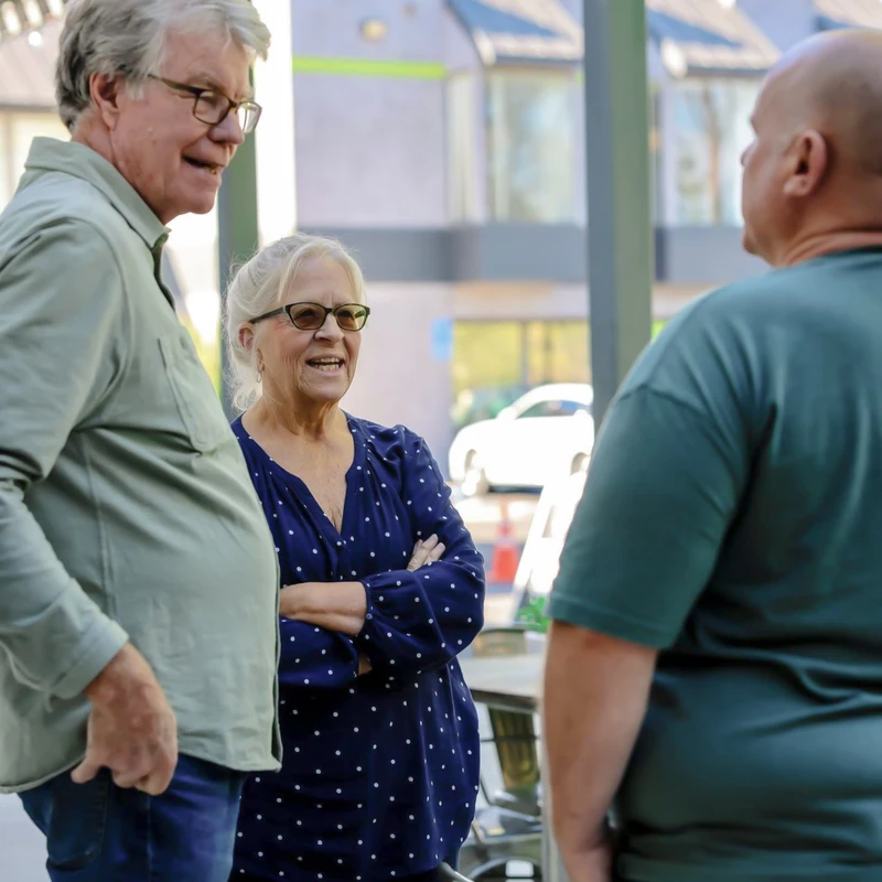 A woman with white hair and a man with gray hair listen as they chat with another man on a sunny patio. The woman is smiling with her arms crossed.