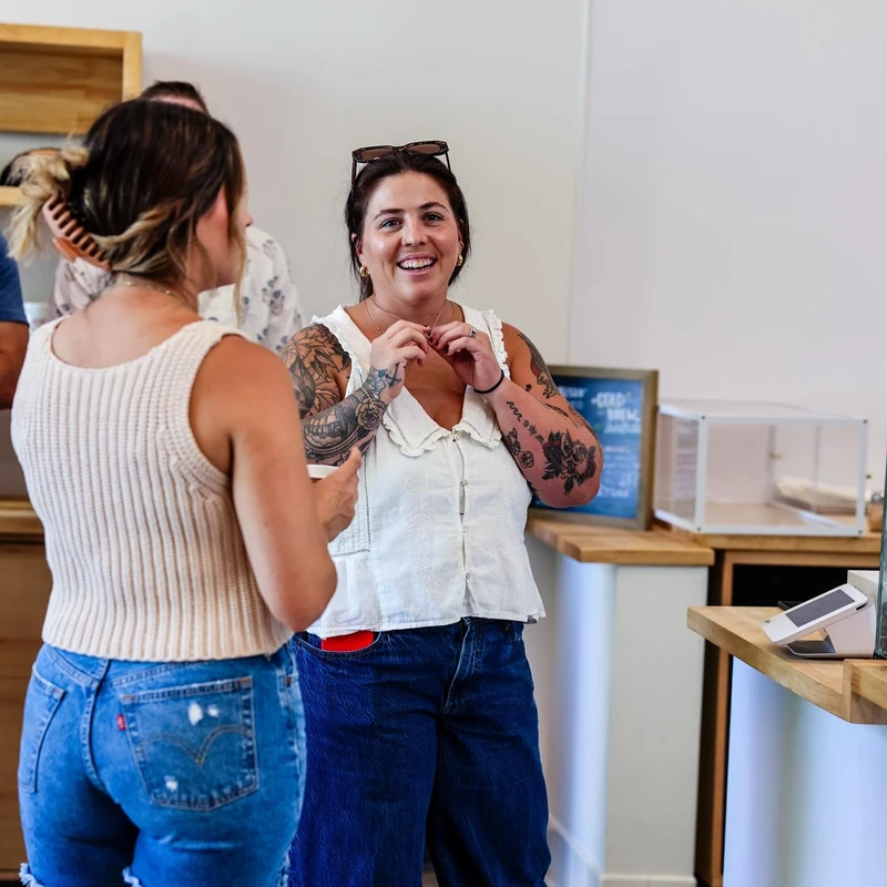 A smiling woman with arm tattoos and sunglasses on her head talks with another woman, seen from behind, while standing at a counter in a brightly lit shop.