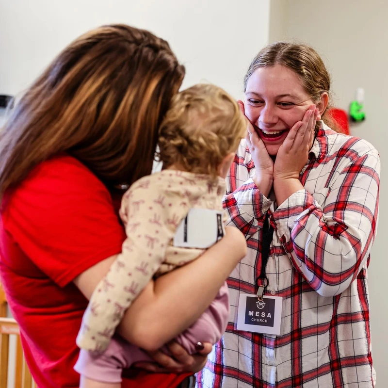 A woman in a plaid shirt reacts with joy, holding her cheeks and smiling, as she greets a baby being held by its mother at a church event.