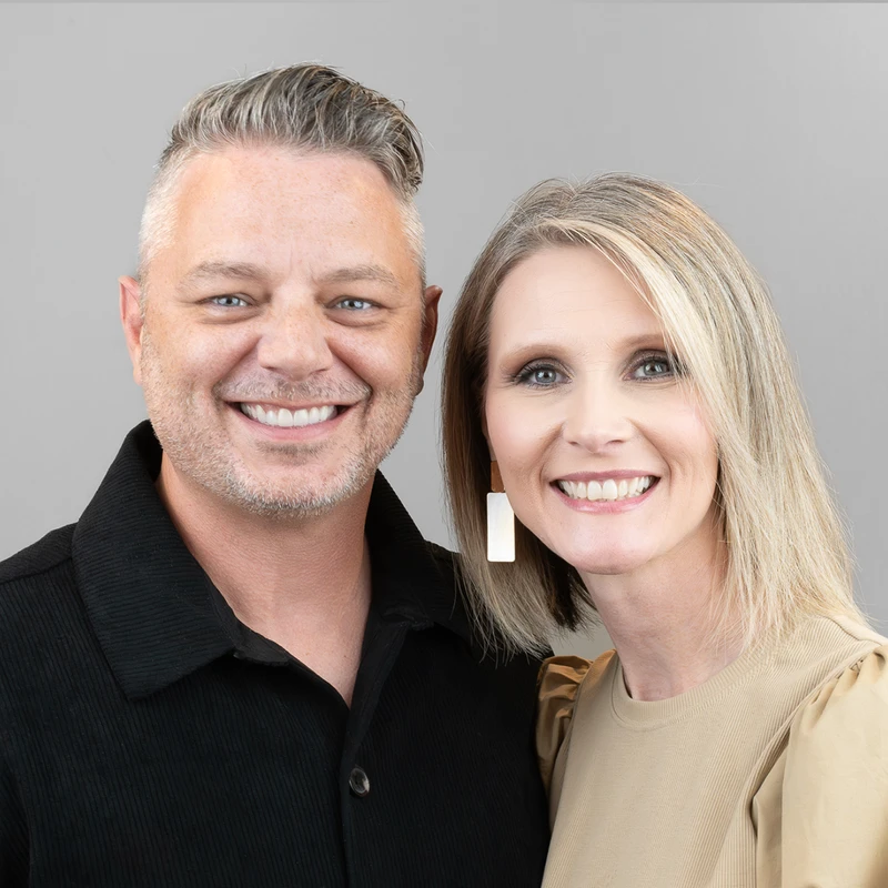 A head and shoulders studio portrait of a smiling man and woman standing side-by-side against a plain gray background.
