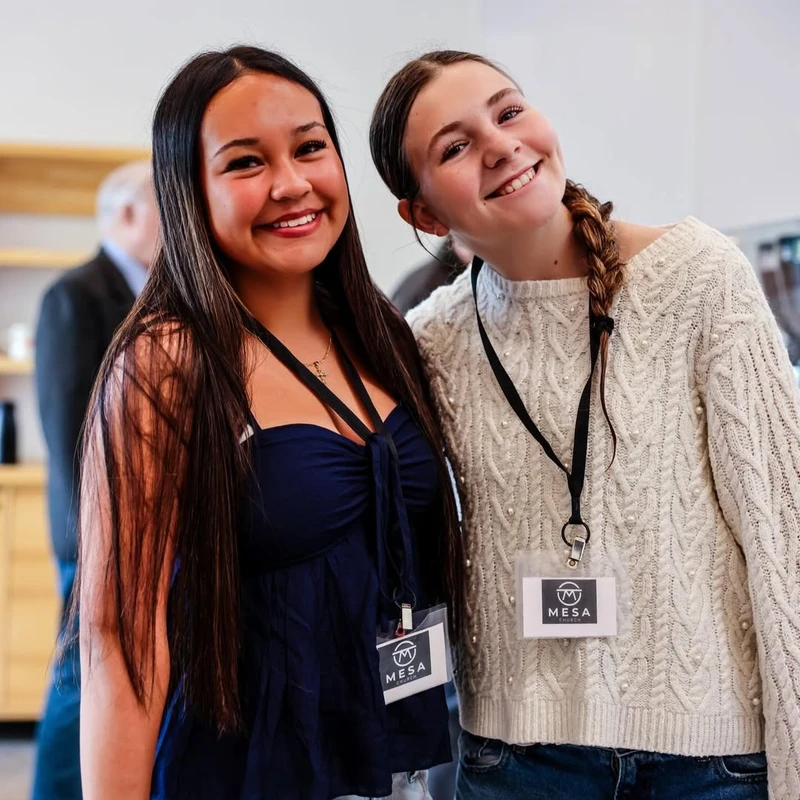 Two young women smile for the camera while at an indoor event. They are both wearing name badges on lanyards with the MESA logo.