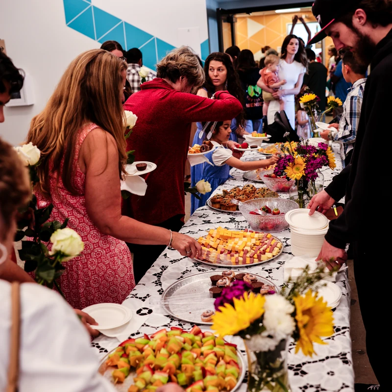 A crowd of people serves themselves from a long buffet table filled with appetizers and decorated with flowers during an indoor social event.