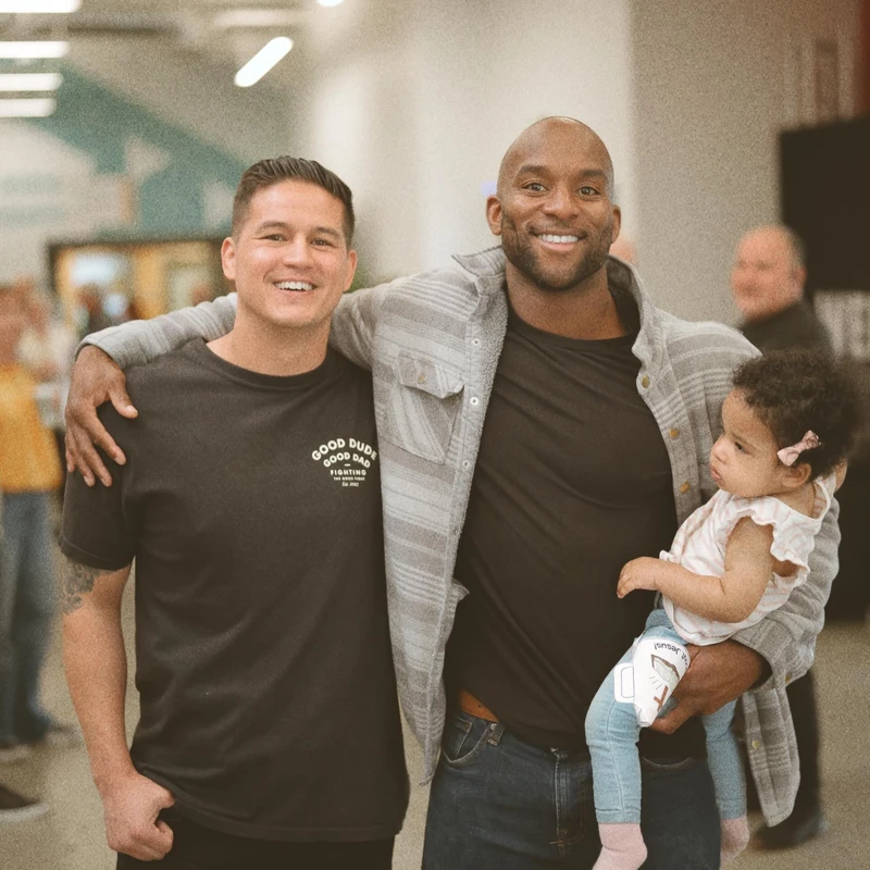 Two men smile for the camera. One man has his arm around the other, who is holding a small child. They are standing together in a well-lit indoor hallway.