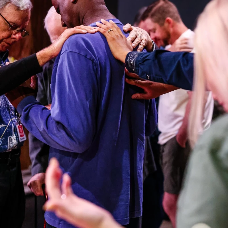 A diverse group of people lay their hands on the shoulders of a man in a blue sweatshirt, bowing their heads in a moment of communal prayer and support.