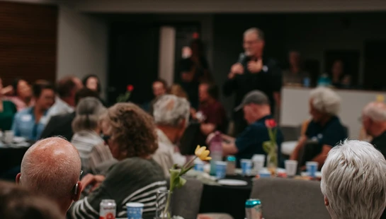 An over-the-shoulder view of people sitting at tables in a room, looking towards a man in the background speaking into a microphone.