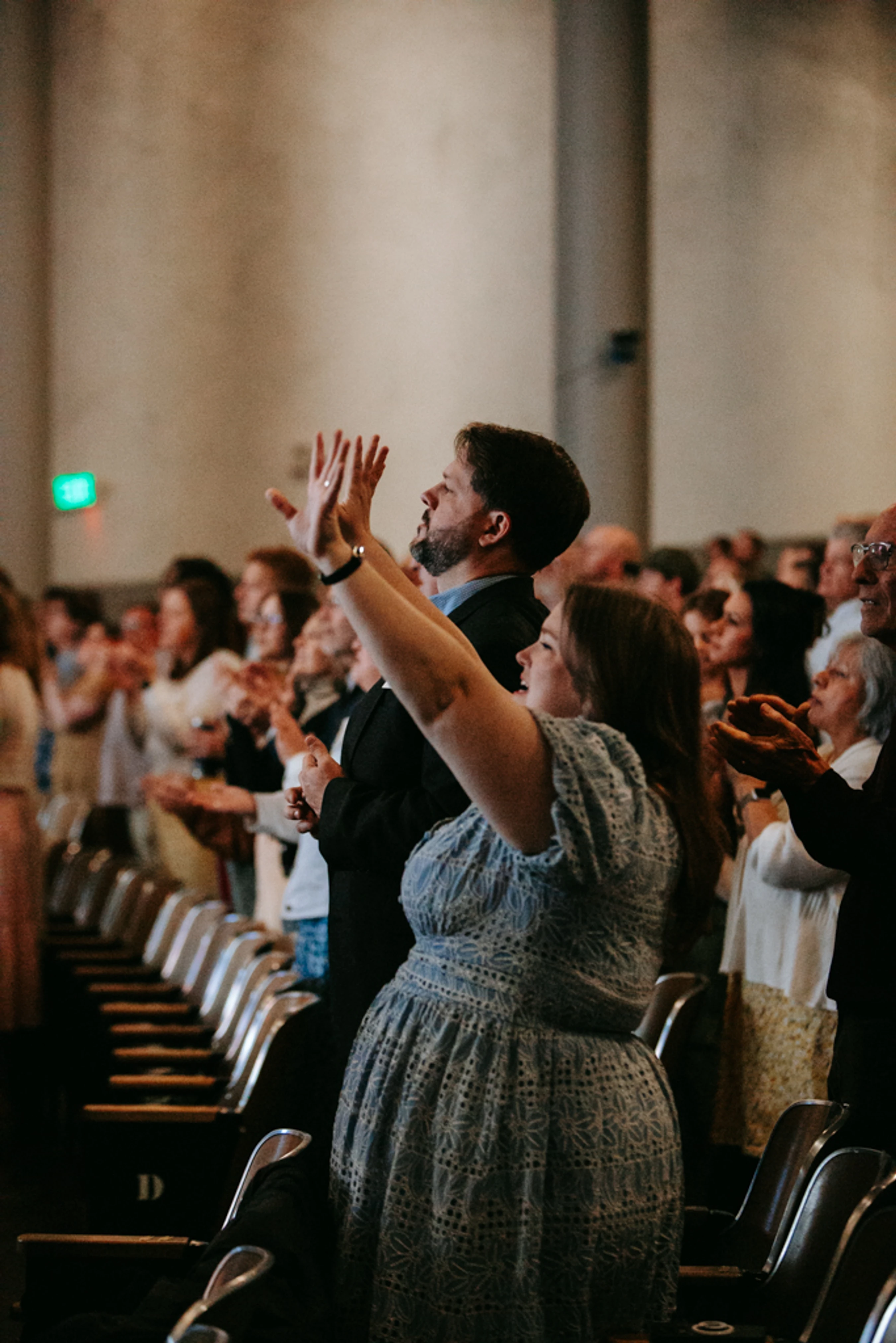 A man and woman in the foreground of a crowd stand with their hands raised in an act of praise or worship inside an auditorium.