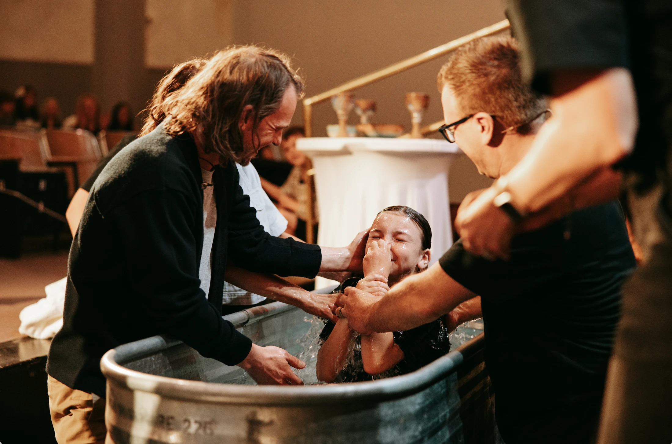 A young person smiles with their eyes closed as two men baptize them in a metal tub filled with water during a church service.