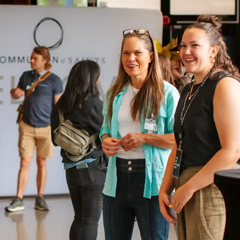 Two women stand together smiling and talking at an indoor event. In the background, other people mingle in front of a wall with a welcome sign.