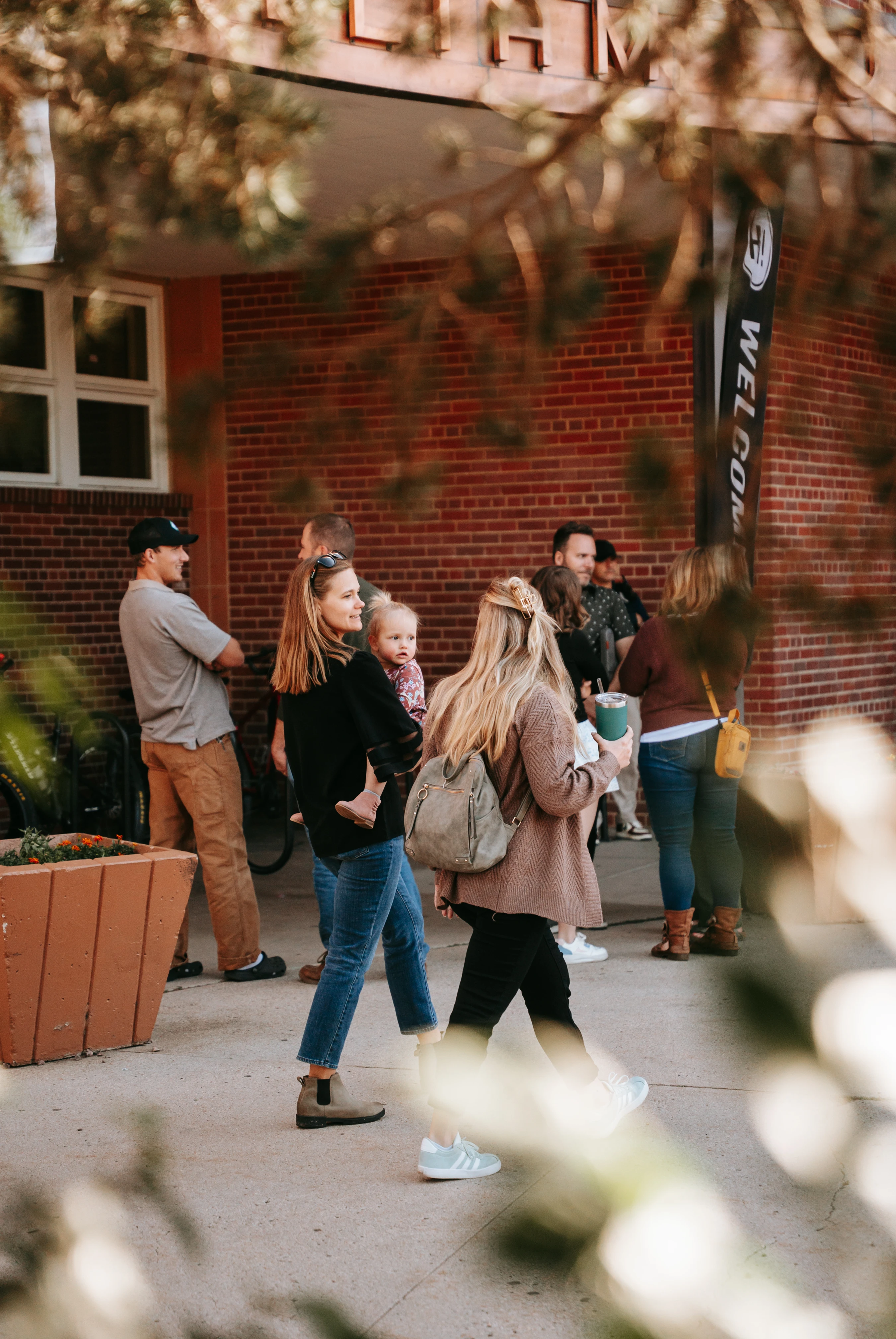 A group of people, including a woman holding a toddler, stand and walk on a sidewalk next to a brick building with a 'WELCOME' flag hanging nearby.