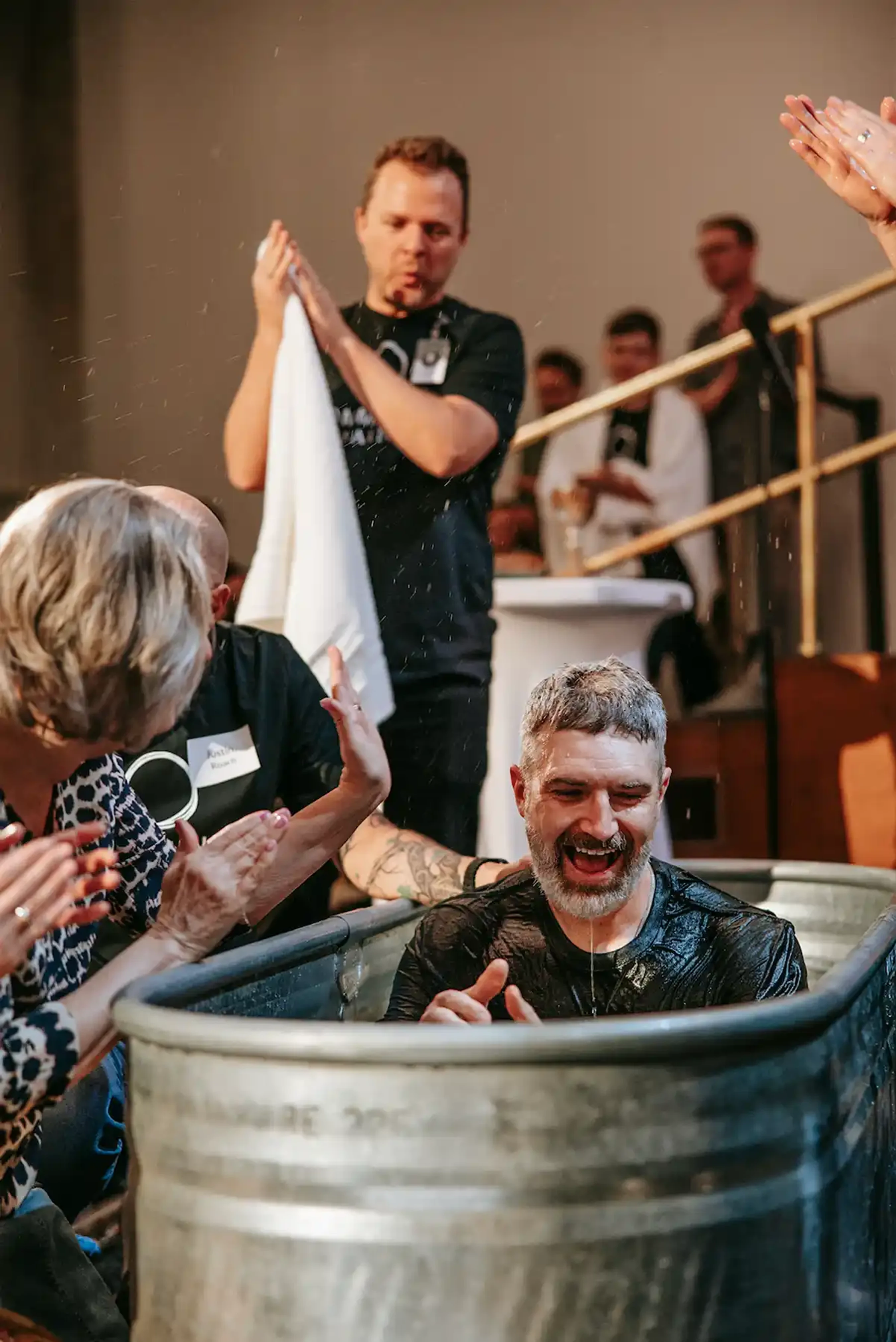 A man with a grey beard laughs joyfully, soaking wet in a metal tub, having just been baptized as people around him clap and celebrate.