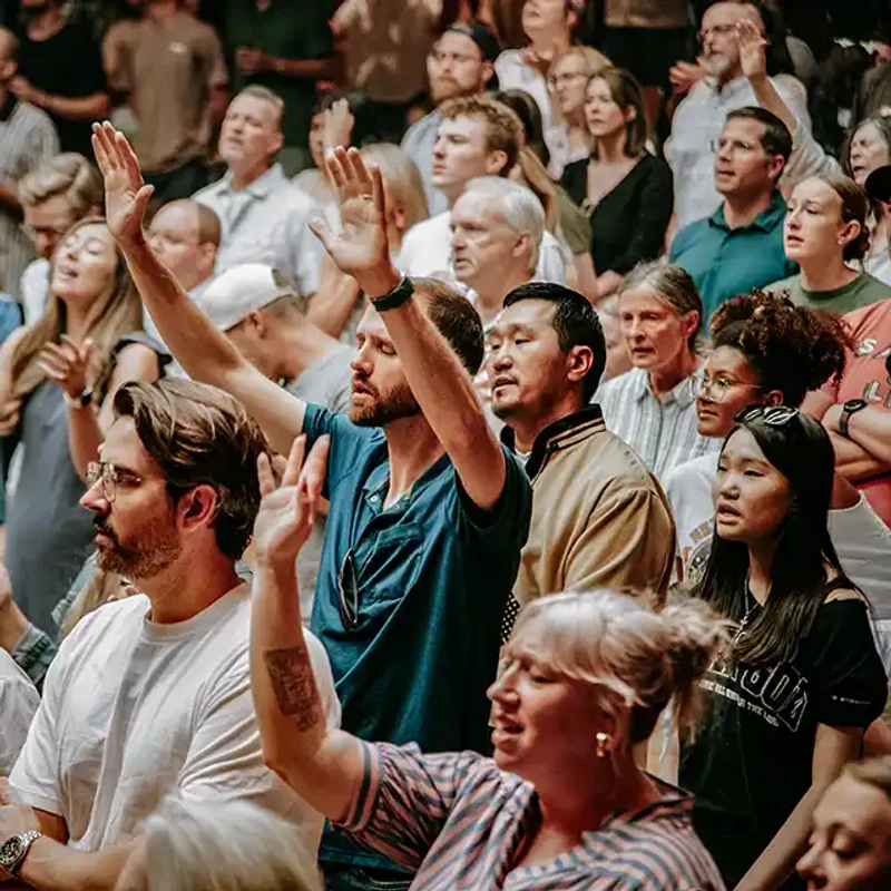 A diverse crowd of people stands together indoors. Many have their hands raised and faces turned upward in an act of communal worship or praise.