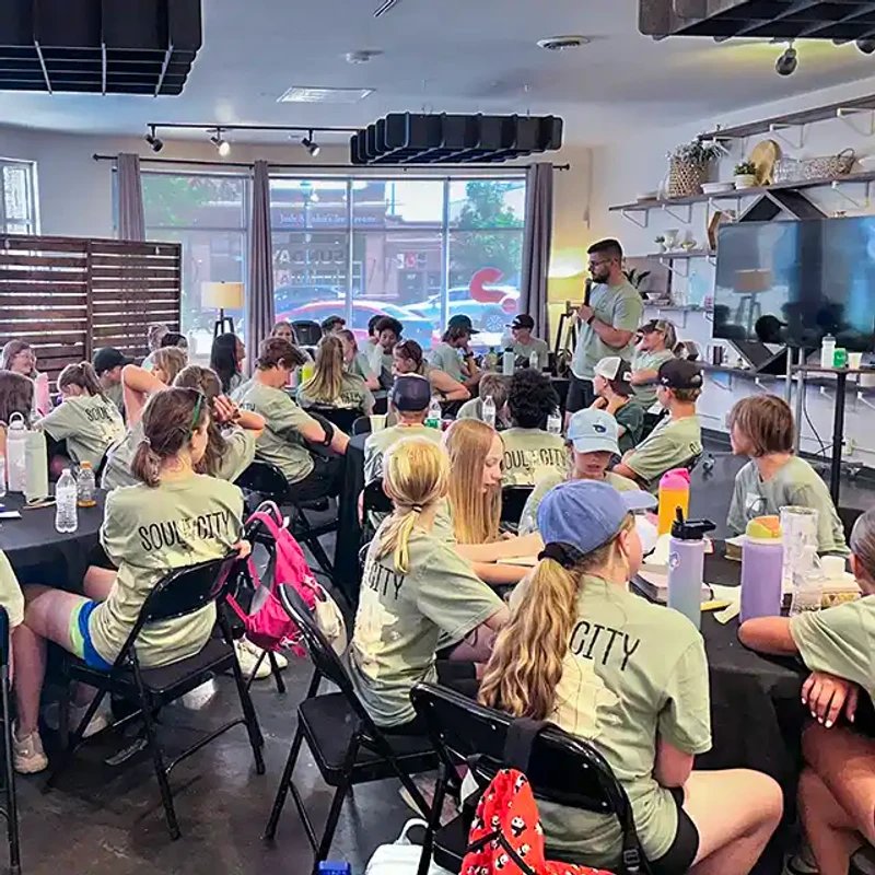 A group of young people in matching 'Soul City' t-shirts sit at tables listening to a man speaking at the front of a room during a youth event.