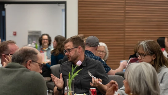 A man with glasses in a grey shirt talks with others at a dinner table during an event. He gestures with his hands while holding a fork.