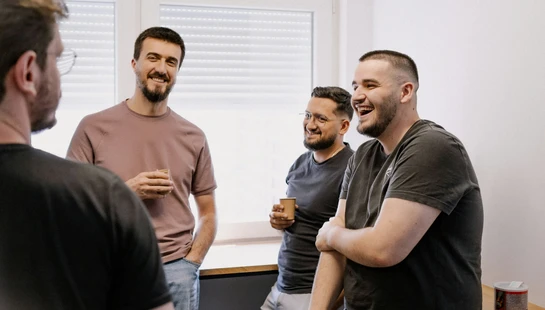 Four casually dressed men stand together in an office, smiling and laughing while holding coffee cups during a break.